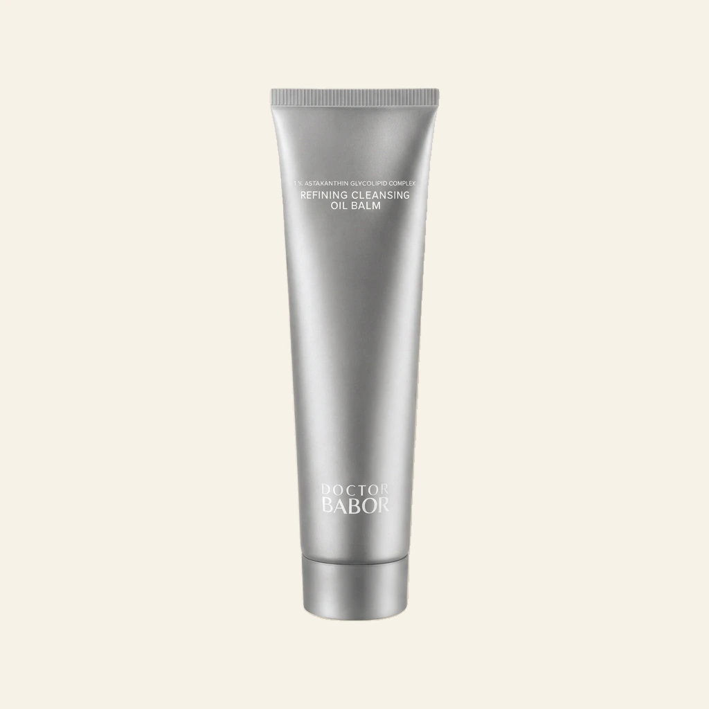 DOCTOR BABOR Refining Cleansing Oil Balm in eleganter silberner Tube vor hellem Spa-Hintergrund mit Naturstein, sanften Beige- und Cremetönen, Kerze und grünen Blättern. Hochwertige Beauty-Produktinszenierung mit modernem Clean-Luxury-Look und entspannender Wellness-Atmosphäre
